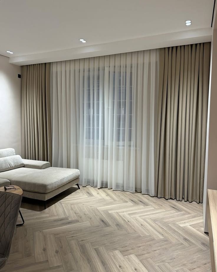 Beige and sheer white curtains hanging in a modern living room with herringbone floors.